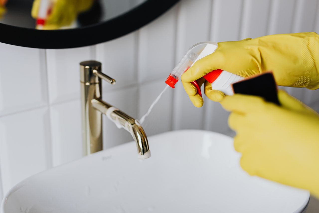 Vue d’en haut d’un agent d’entretien anonyme portant des gants en caoutchouc jaunes et pulvérisant un nettoyant sur un robinet d’eau chromé dans une salle de bain